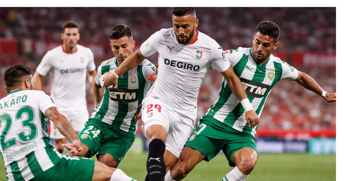“Sevilla player in white kit shields the ball while being challenged by three Elche players in green and white during a high-intensity La Liga match inside a packed stadium under floodlights.”