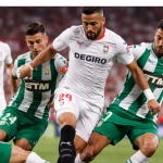 “Sevilla player in white kit shields the ball while being challenged by three Elche players in green and white during a high-intensity La Liga match inside a packed stadium under floodlights.”